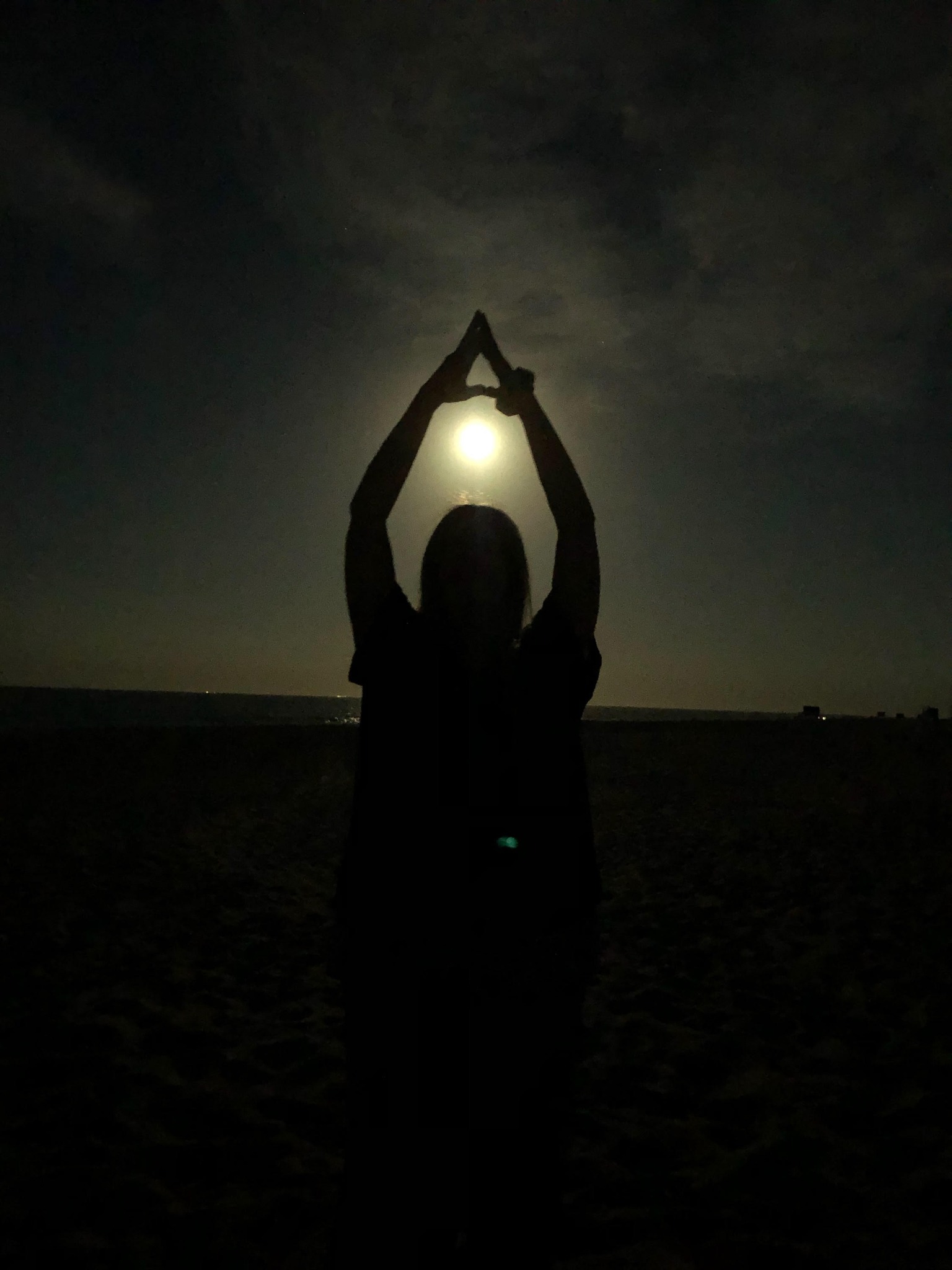 Silhouette of a person forming a triangle with their arms around the moon at night on a beach